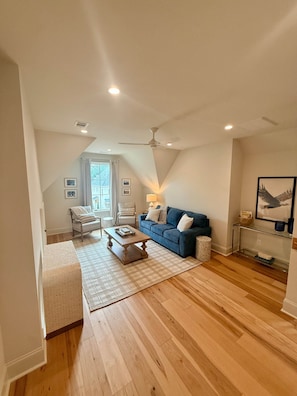 Ein großzügiges Wohnzimmer mit hoher Decke, Deckenventilator, blauem Sofa, Holztisch und einem großen Fenster mit Vorhängen.