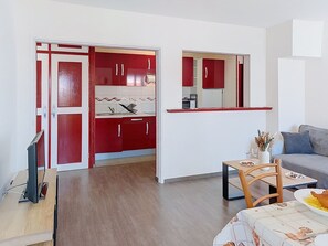 A modern living room with a grey sofa, a dining table with a vase and glasses, and a kitchen area with red cabinets and a microwave. - Les Dunes by Interhome (Mimizan)