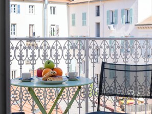 Ein Balkon mit Tisch, gedeckt mit Früchten, einer Tasse und einem Gebäck, mit Blick auf eine Reihe von Gebäuden mit Balkonen.