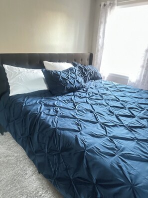 A bed with a dark blue quilt and two pillows, a tufted headboard, and a window with curtains.