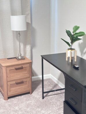 A room with a wooden nightstand, a desk with drawers, a lamp, and a potted plant.