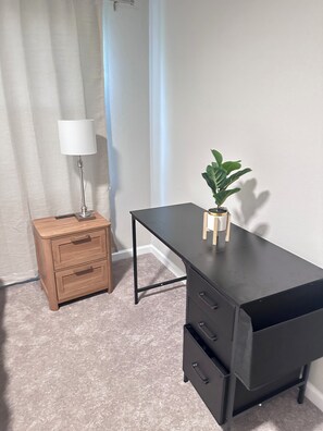 Ein Zimmer mit Nachttisch aus Holz, einem schwarzen Schreibtisch, einer Lampe und einem Topf mit Pflanze.