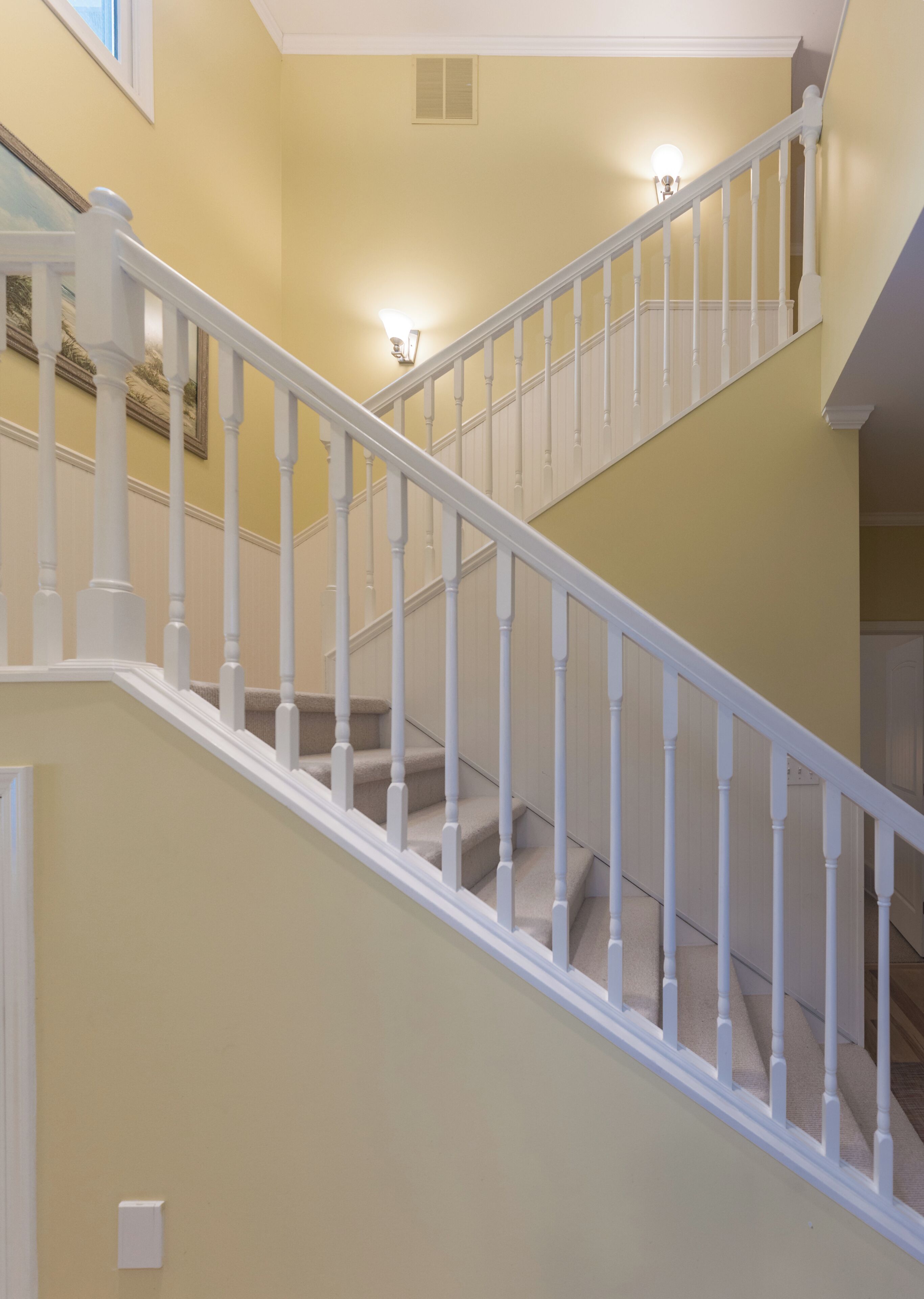 Un escalier avec des balustrades blanches et une moquette beige, menant à un palier où se trouve un luminaire.