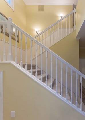 Un escalier avec des balustrades blanches et une moquette beige, menant à un palier où se trouve un luminaire.