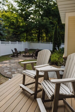 Une terrasse dotée de chaises Adirondack et d’un foyer, entourée d’une clôture blanche et de verdure.