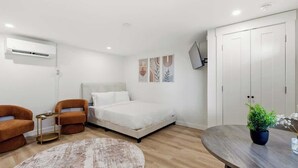 Een moderne slaapkamer met een bed, twee fauteuils, een tafeltje, een rond vloerkleed, een kamerplant en een kledingkast.