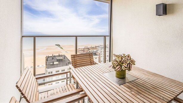 Een balkon met een houten tafel, een bloempot en uitzicht op een stadslandschap en woestijn.