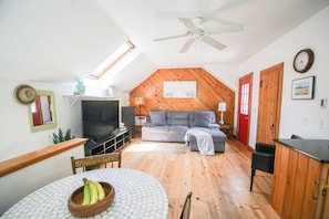 A cozy loft-style room with a wooden ceiling, a gray sofa, a television, and a dining area with a table and chairs.
