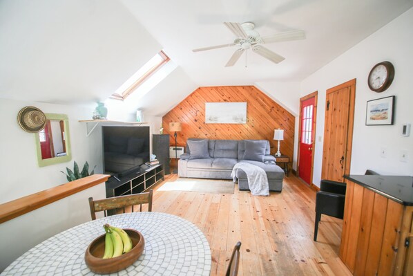 A cozy loft-style room with a wooden ceiling, a gray sofa, a television, and a dining area with a table and chairs.