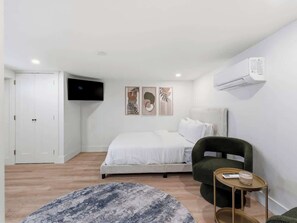 A bedroom with a bed, a chair, a small table, and a mounted air conditioner.