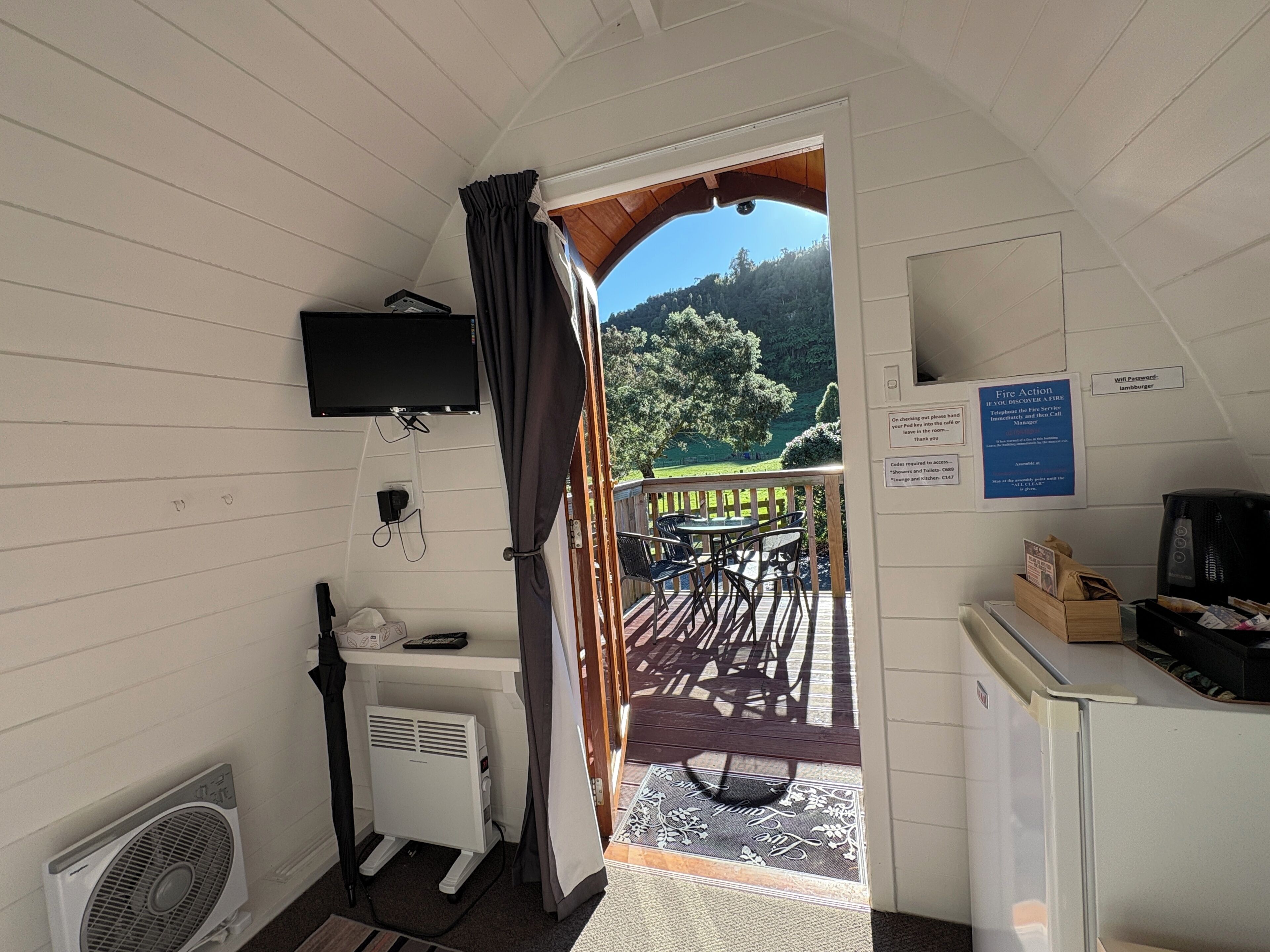 A room with a television mounted on the wall, a small desk with a computer, a heater, and a door leading to an outdoor area with a table and chairs.