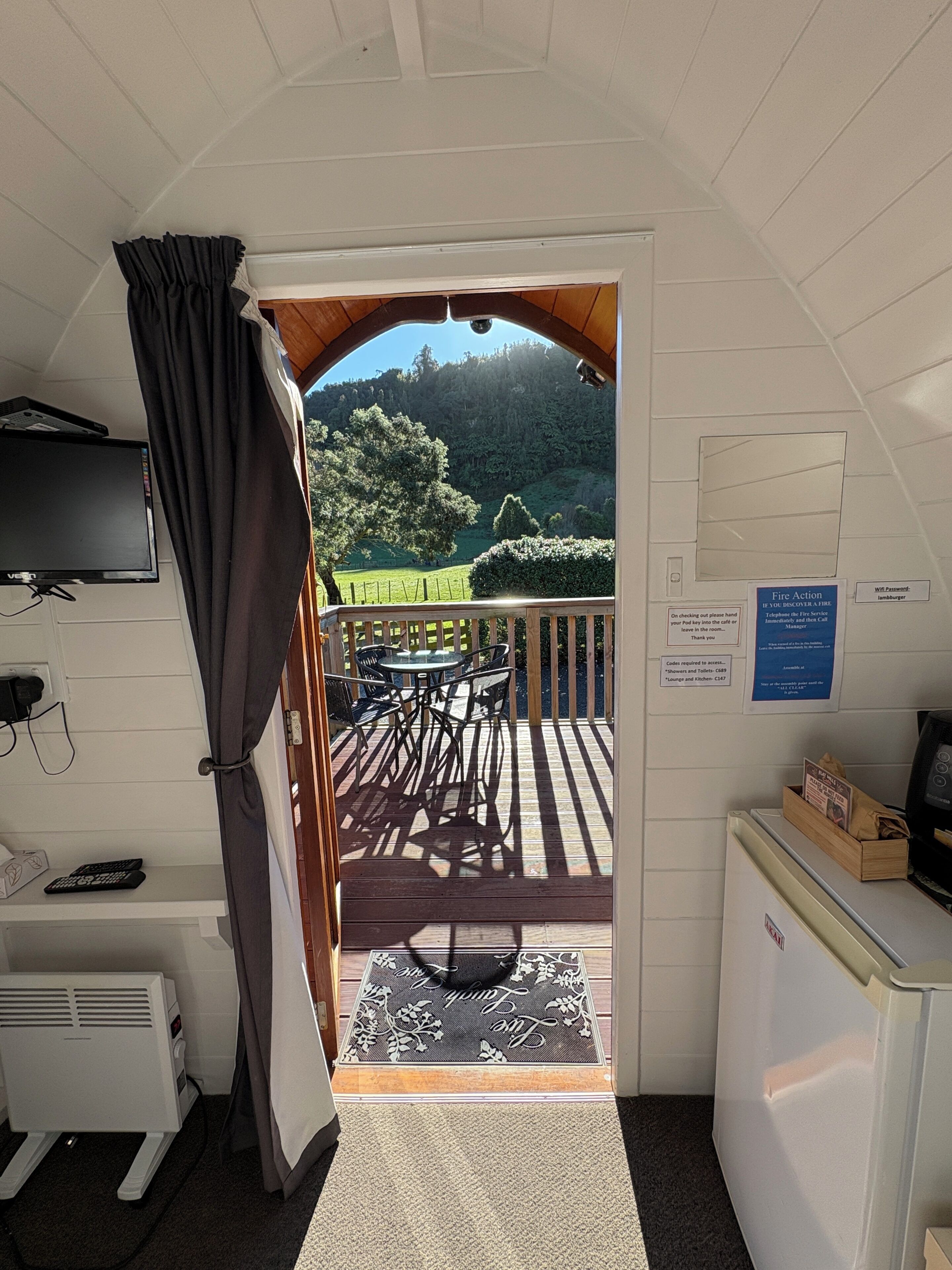 A room with a TV, a desk, a computer, a window with curtains, and a view of a deck with outdoor furniture.
