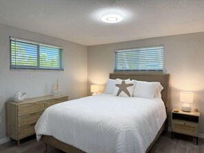 A bedroom with a bed, two bedside tables, a nightstand, a wall-mounted lamp, a decorative sailboat, and two windows with blinds. - 2BR Coastal Beach & Boating Escape (Englewood)