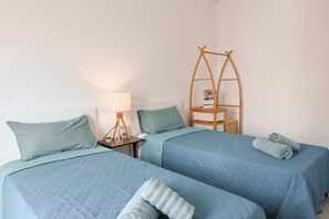 A room with two beds, each with a blue quilted bedspread and matching pillows. There is a wooden ladder shelf against the wall, a bedside table with a lamp, and a small basket. - Yacht Marina View - Modern 3 bdrs Apart M, Msida, Malta (Msida)