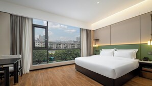 A modern hotel room with a large bed, a desk, and a view of a cityscape through a large window. - Baihe yiting jiudian (Foshan)