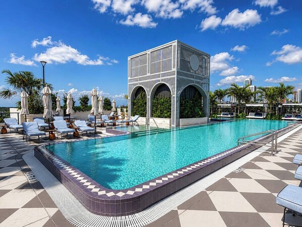 A swimming pool with lounge chairs and umbrellas, a covered seating area, and a modern building with arches.