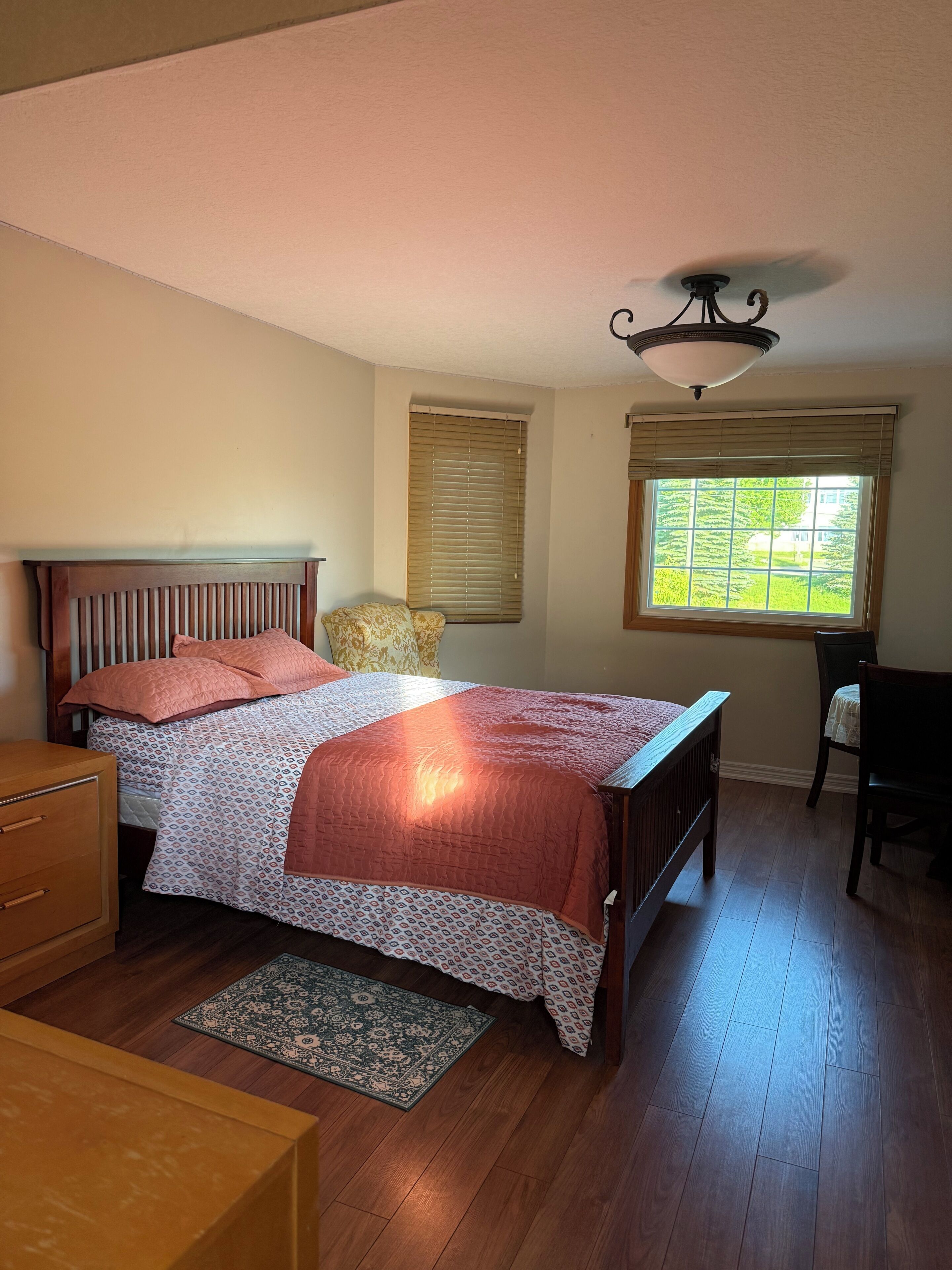 A bedroom with a wooden bed, a nightstand, a chair, a window with blinds, and a ceiling fan.