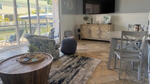 Un salón con televisor, una mesa de comedor, sillas, un sofá, una mesa baja y vista a un cuerpo de agua exterior.