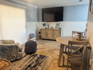 Ein Wohnzimmer mit einer Couch, einem Hocker und einem Esstisch mit Stühlen. Ein Fernseher ist an der Wand über der Couch angebracht.