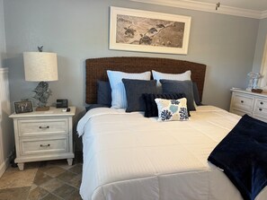 Ein ordentlich eingerichtetes Schlafzimmer mit einem Bett, Kissen, einem Nachttisch, einer Lampe und einem gerahmten Bild an der Wand.