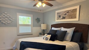 Un dormitorio con cama, ventilador de techo, ventana con persianas, lámpara de pared y un cuadro enmarcado.