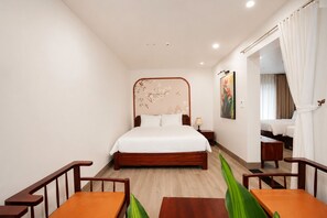 A hotel room with a large bed, a desk, two chairs, a small table, a nightstand, a lamp, a painting, and a window with curtains.