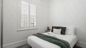 A bedroom with a bed, a window with blinds, a bedside table with a lamp, and a nightstand. - The Leichhardt Hideaway on Francis St (Leichhardt)