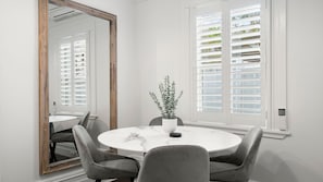 Ein Essbereich mit einem runden Tisch, vier Stühlen, einem Blumentopf, einem großen Spiegel und einem Fenster mit Fensterläden.