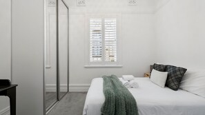 Une chambre à coucher avec un lit, une fenêtre avec des stores, un miroir et une table de chevet.
