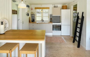 Eine Küche mit Holztisch, zwei Korbstühlen, einem Kühlschrank, einem Backofen und einem Fenster mit Jalousien.