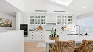 A modern kitchen with white cabinetry, a built-in oven, and a large island with seating. - Glass House Retreat at Forresters (Forresters Beach)