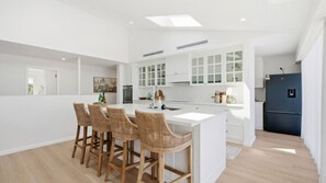 A modern kitchen with a central island, bar stools, white cabinetry, and a dark-colored refrigerator. - Glass House Retreat at Forresters (Forresters Beach)
