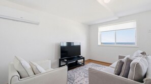 A living room with a flat-screen TV on a stand, two sofas, a patterned rug, and a window with a view.