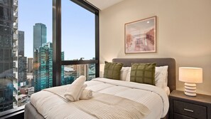Una habitación de hotel con un ventanal grande, una cama con ropa blanca, una mesita de noche con lámpara y un cuadro en la pared.