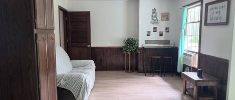 A room with a bed, a wooden wardrobe, a kitchenette with a bar area, and a window with curtains.