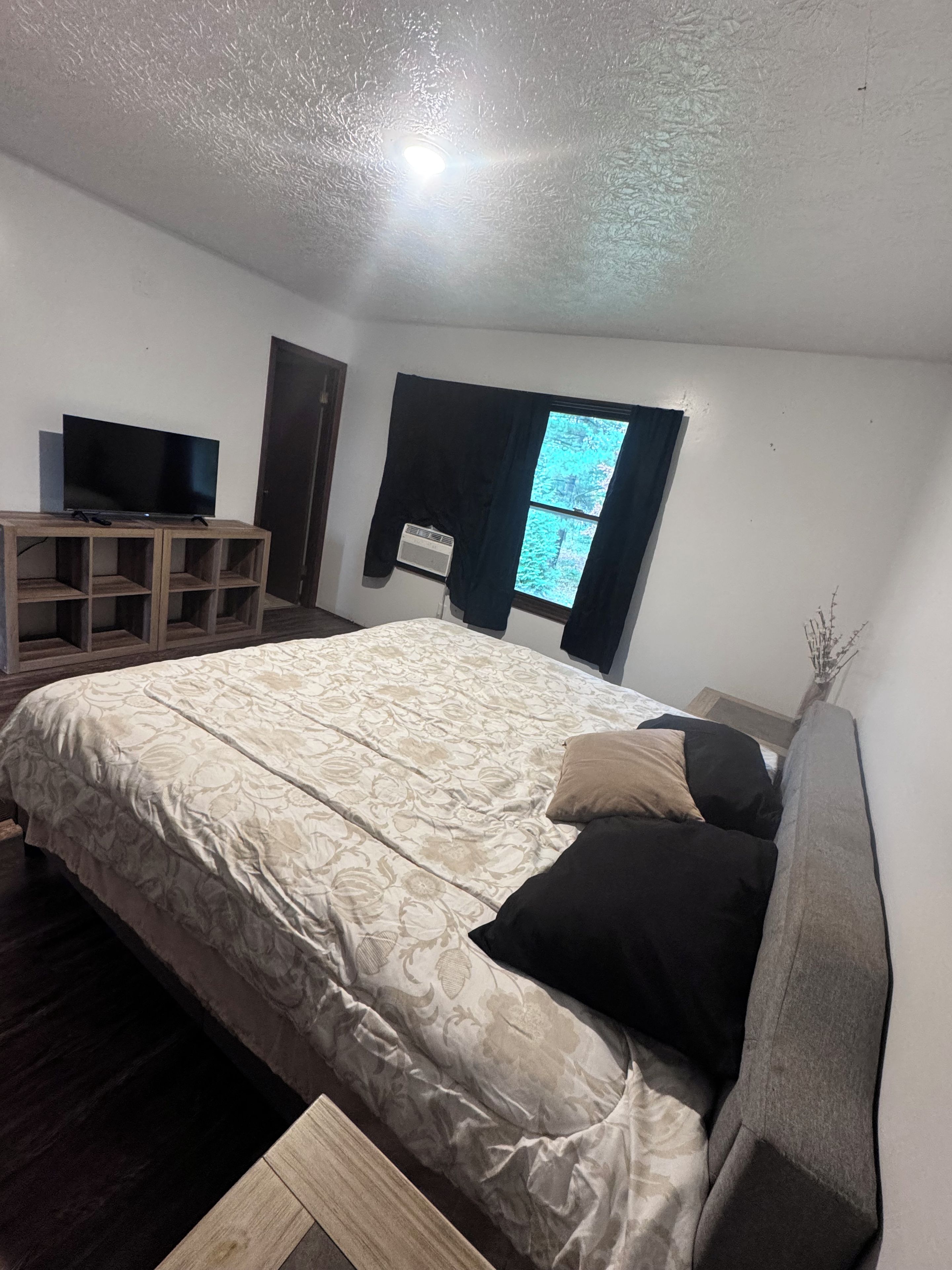 A bedroom with a bed, a TV on a wooden stand, a window with curtains, and a radiator.