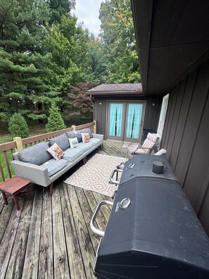 A deck with a grill, a sofa with cushions, and a small building with a door and curtains.