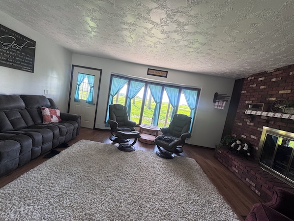 A living room with a dark leather sofa, two armchairs, a white rug, a fireplace, and a view of a green landscape through large windows.
