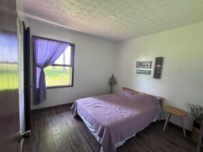 A bedroom with a bed, a window with curtains, a small bedside table, a potted plant, and a wall-mounted sign.