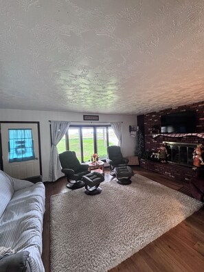 Een woonkamer met een groot raam, twee fauteuils, een bank en een open haard met decoraties.