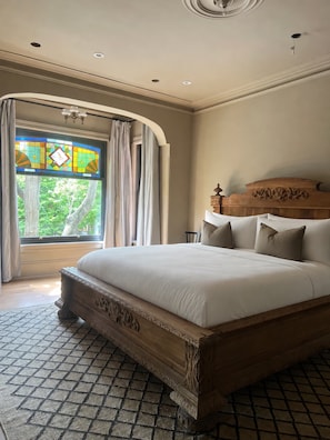 A bedroom with a large bed, a wooden headboard, a stained glass window, and a view of greenery outside.