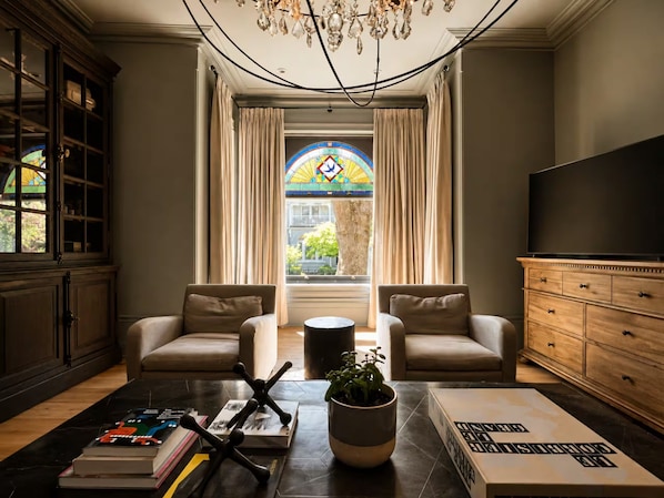 A living room with two armchairs, a coffee table, a bookshelf, a TV, and a stained glass window.