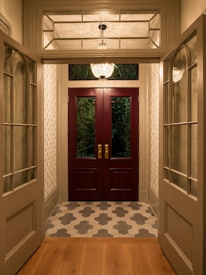 A hallway with a double door, patterned wallpaper, hexagonal patterned flooring, and a chandelier.