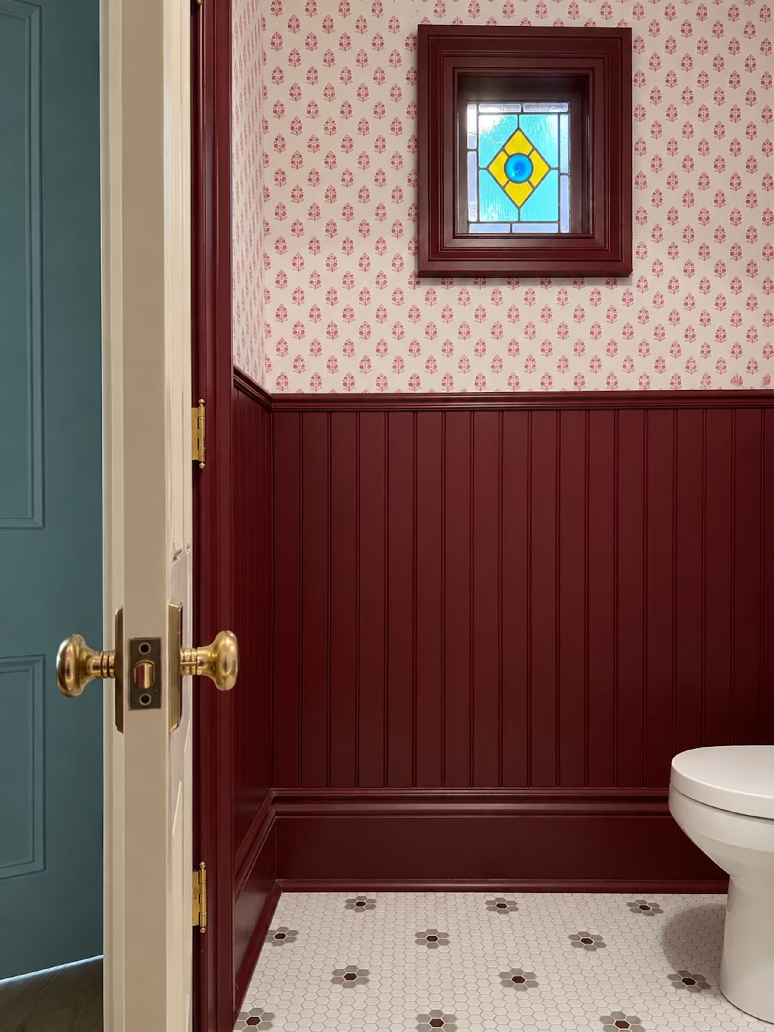 A bathroom with a white toilet, a patterned wall, a small window with a diamond-shaped stained glass, and a tiled floor.
