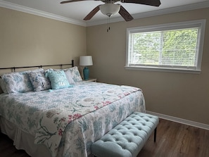 A bedroom with a bed, a ceiling fan, a window with blinds, a nightstand with a lamp, and a bench.