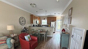 A living room with a red sofa, a dining area with a table and chairs, a kitchen with wooden cabinets and a refrigerator, and a door leading outside. - No Egrets: 2 BR Condo Gulf Views, Pool, Walk to Beach, Golf Cart District (Long Beach)