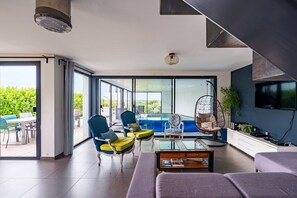 Uma sala de estar moderna com uma porta de vidro que dá para uma área externa, um sofá, cadeiras, uma mesa de centro e uma TV fixada na parede.