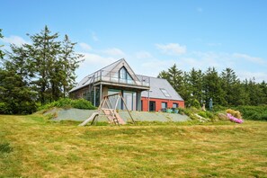 Et moderne hus med en stor glasbalkon, et gyngestativ af træ og en rød bygning i baggrunden.