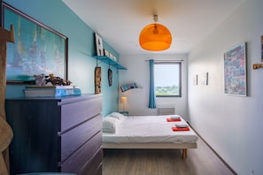 Un dormitorio con cama, cómoda, estanterías con libros y objetos decorativos, una ventana con cortinas y una lámpara colgante.