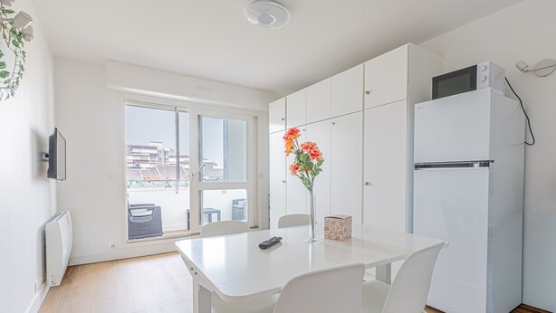 Eine moderne Küche mit weißen Schränken, einem weißen Kühlschrank, einem Esstisch mit Stühlen und einem Fenster mit Blick auf Gebäude.